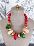 A chunky statement necklace with a mix of red, gold, green, and pale pink polished beads, displayed on a beige fabric mannequin bust.