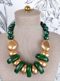 A statement necklace made of alternating polished green beads and chunky gold disc-like beads, displayed on a beige fabric mannequin bust.
