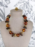 A statement necklace made of polished amber and dark brown glass beads with gold spacer accents, displayed on a beige fabric mannequin bust against a black and white floral wallpaper background.