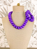 A chunky purple beaded statement necklace set with stretchy bracelet displayed on a beige fabric mannequin bust, set against a light wallpaper with botanical line drawings.