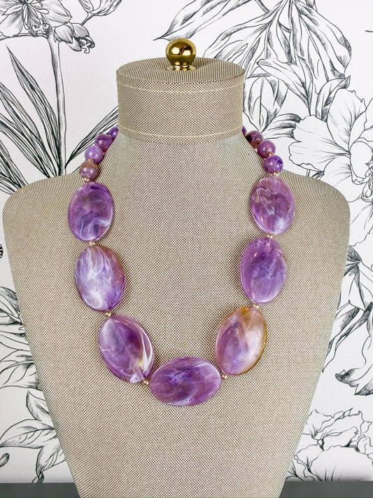 A purple marbled stone bead necklace displayed on a beige fabric-covered jewelry bust against a black-and-white floral background.