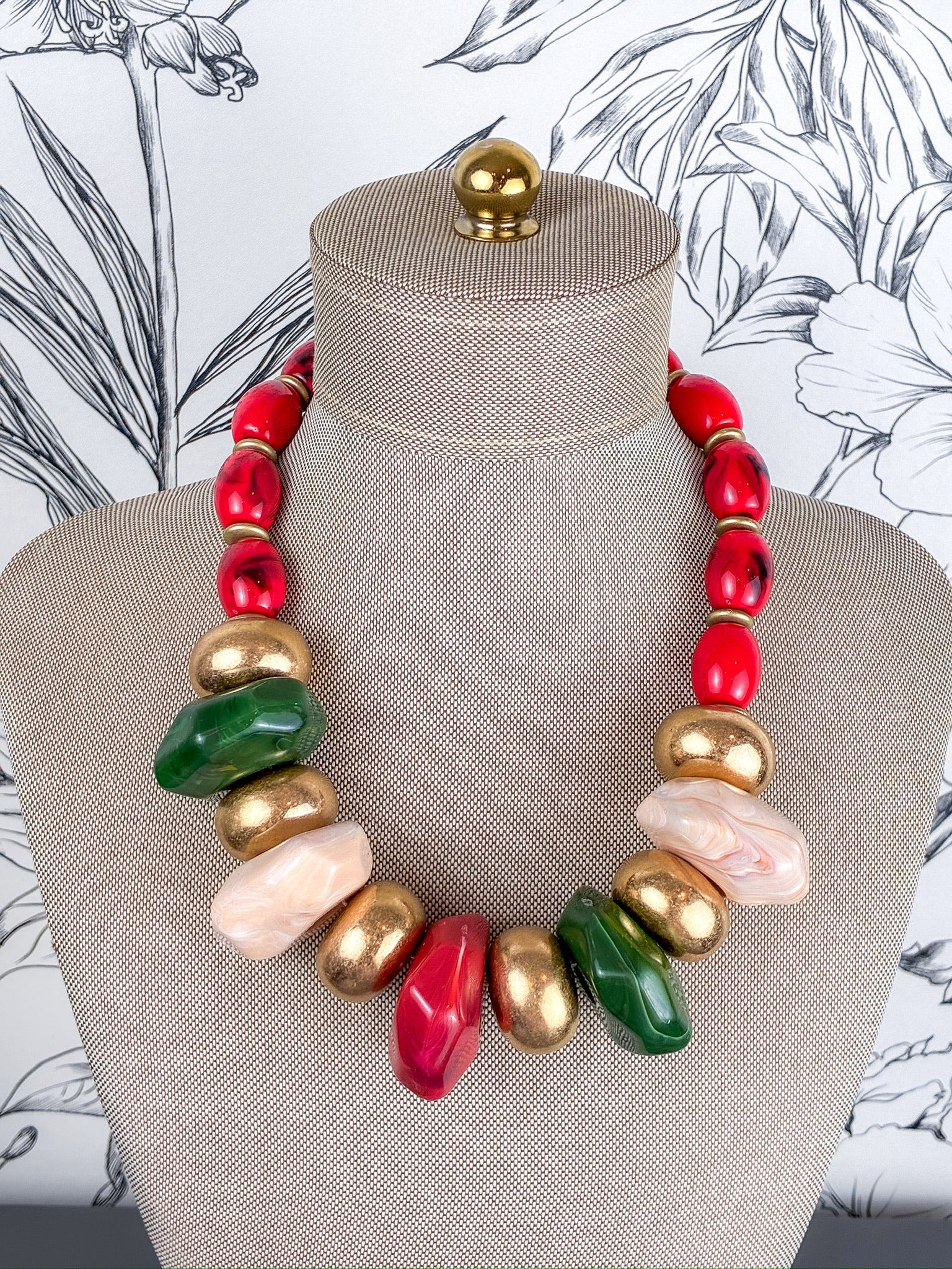 A chunky statement necklace with a mix of red, gold, green, and pale pink polished beads, displayed on a beige fabric mannequin bust.