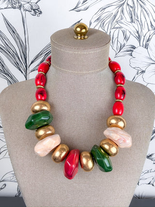 A chunky statement necklace with a mix of red, gold, green, and pale pink polished beads, displayed on a beige fabric mannequin bust.