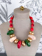 A chunky statement necklace with a mix of red, gold, green, and pale pink polished beads, displayed on a beige fabric mannequin bust.