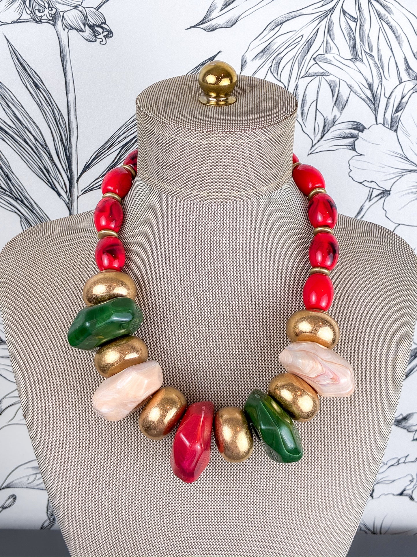 A chunky statement necklace with a mix of red, gold, green, and pale pink polished beads, displayed on a beige fabric mannequin bust.