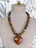 A beaded necklace displayed on a beige fabric mannequin bust. The necklace features alternating strands of polished brown and black tiger's eye beads with a large, smooth, faceted brown heart-shaped pendant at the center.