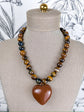 A beaded necklace displayed on a beige fabric mannequin bust. The necklace features alternating strands of polished brown and black tiger's eye beads with a large, smooth, faceted brown heart-shaped pendant at the center.