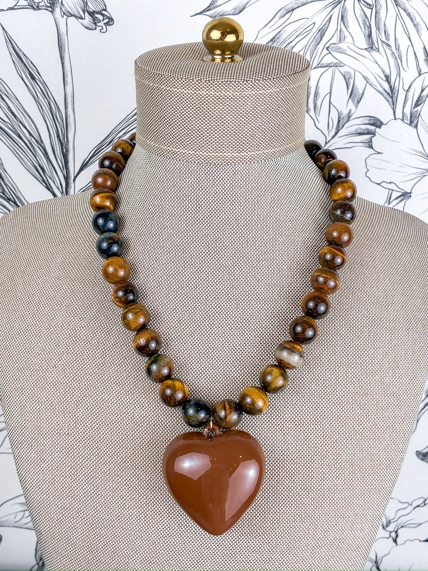 A beaded necklace displayed on a beige fabric mannequin bust. The necklace features alternating strands of polished brown and black tiger's eye beads with a large, smooth, faceted brown heart-shaped pendant at the center.