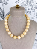 A chunky beaded necklace made of large, polished cream-colored round beads separated by small gold spacers, displayed on a textured beige fabric jewelry mannequin bust.