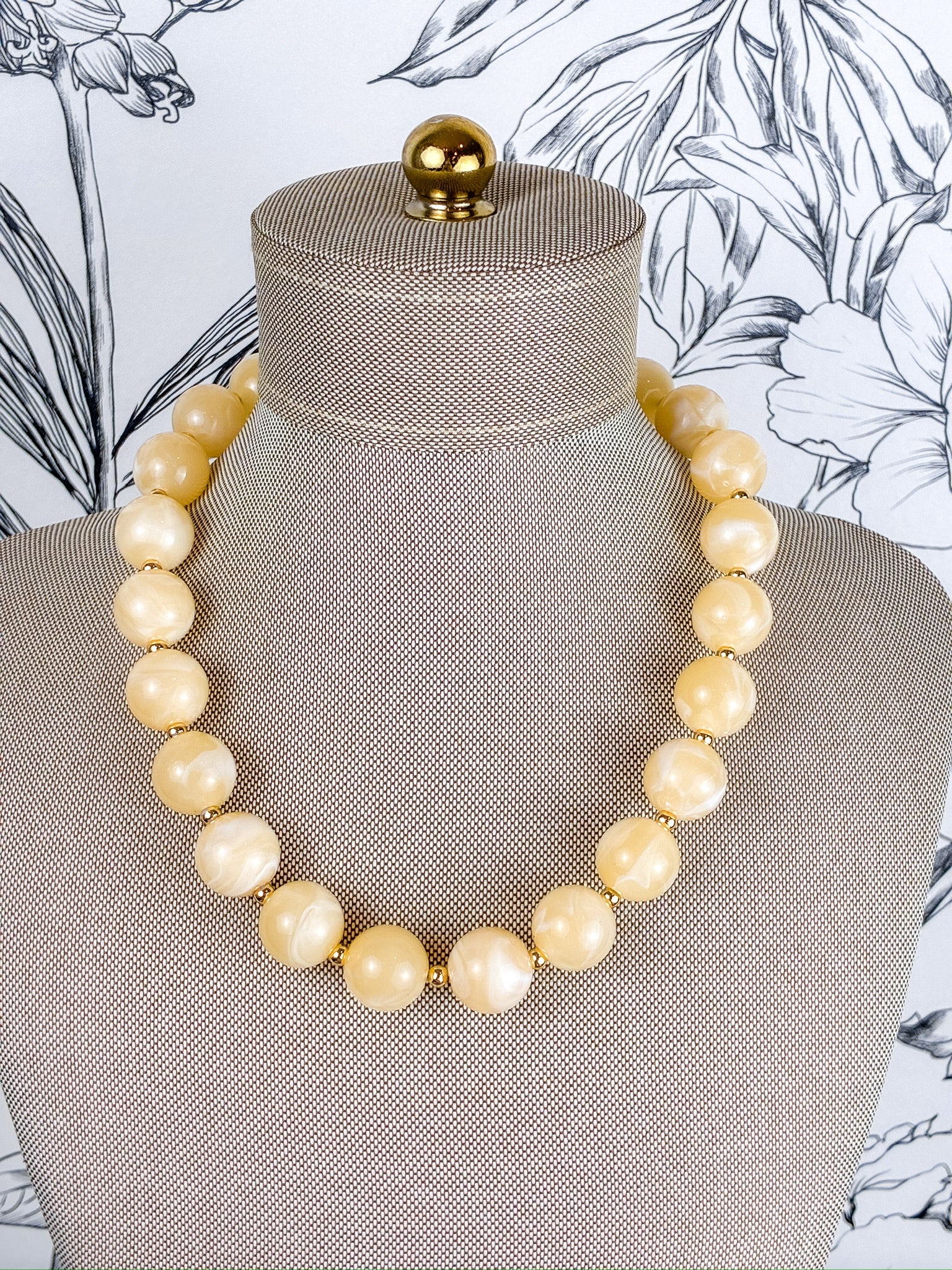 A chunky beaded necklace made of large, polished cream-colored round beads separated by small gold spacers, displayed on a textured beige fabric jewelry mannequin bust.