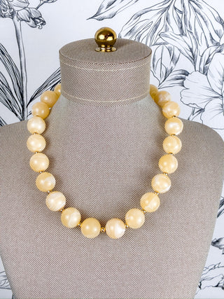 A chunky beaded necklace made of large, polished cream-colored round beads separated by small gold spacers, displayed on a textured beige fabric jewelry mannequin bust.