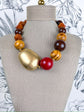 A statement necklace displayed on a beige fabric mannequin bust, featuring a large gold-toned irregular stone, a glossy red round bead, and alternating brown and yellow wood beads.