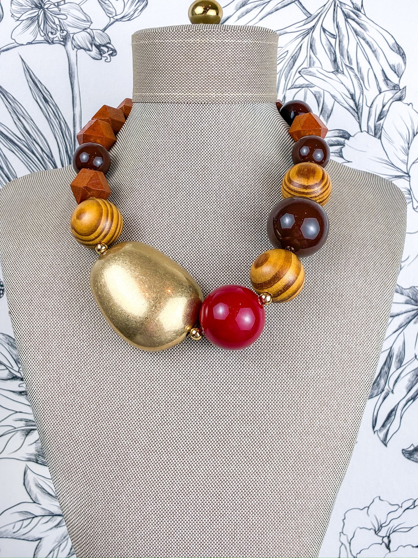 A statement necklace displayed on a beige fabric mannequin bust, featuring a large gold-toned irregular stone, a glossy red round bead, and alternating brown and yellow wood beads.