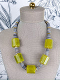 A statement necklace displayed on a beige fabric mannequin bust, featuring large translucent lime green rectangular beads alternating with gray and gold striped spacer beads.