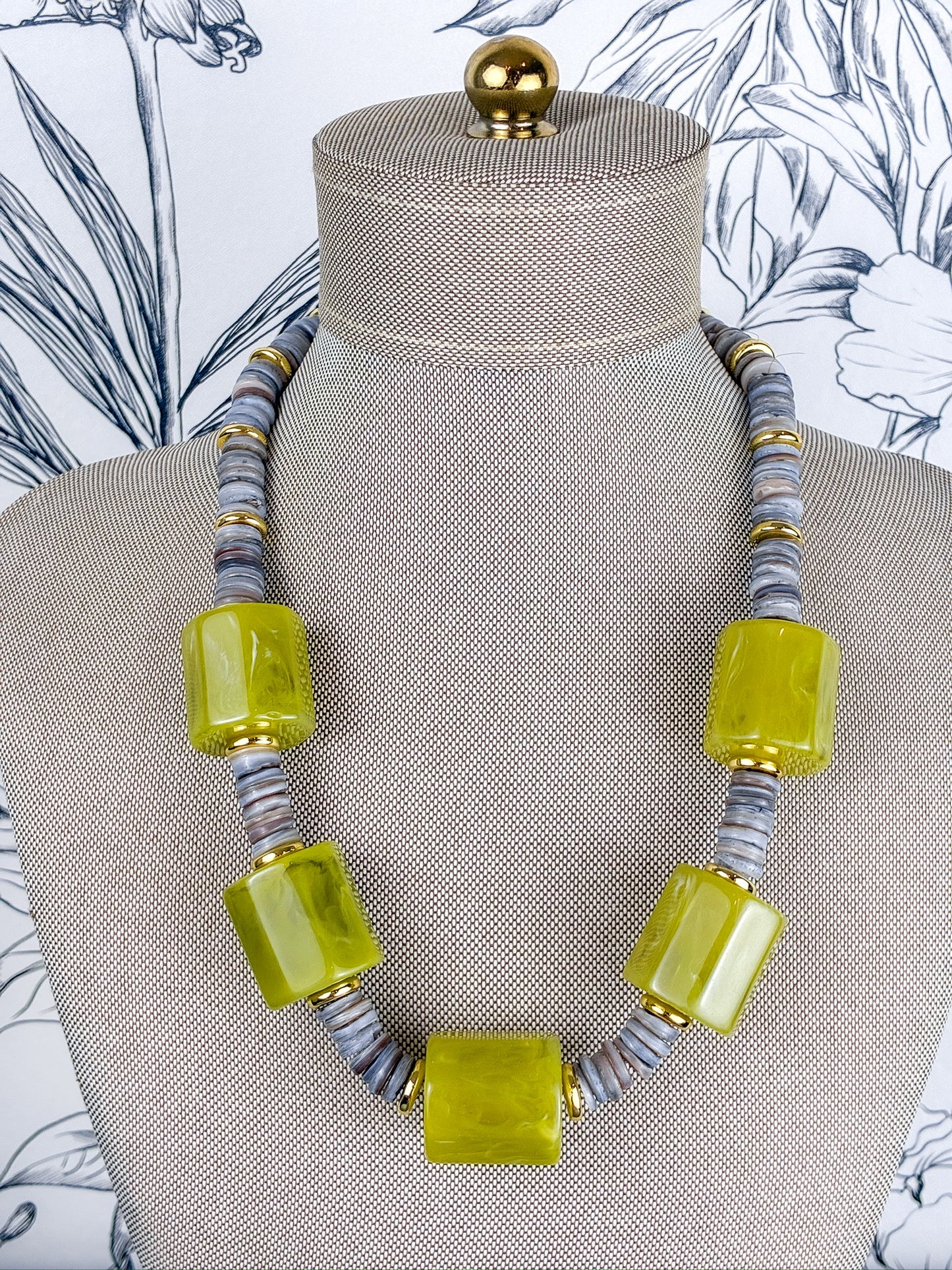 A statement necklace displayed on a beige fabric mannequin bust, featuring large translucent lime green rectangular beads alternating with gray and gold striped spacer beads.