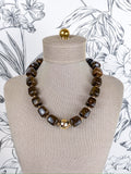A chunky beaded necklace with dark brown tiger's eye stones and a central gold-toned floral accent, displayed on a beige fabric mannequin bust against a black and white floral wallpaper background.