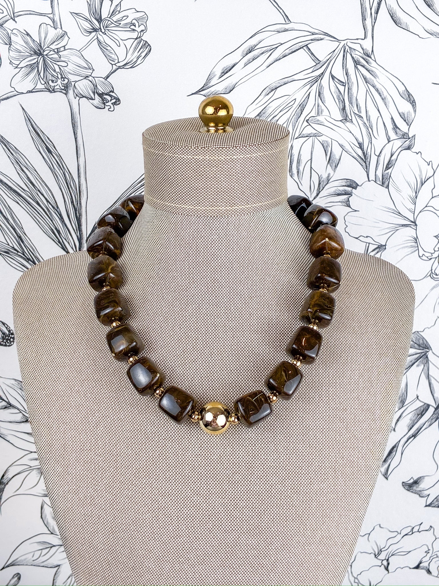 A chunky beaded necklace with dark brown tiger's eye stones and a central gold-toned floral accent, displayed on a beige fabric mannequin bust against a black and white floral wallpaper background.