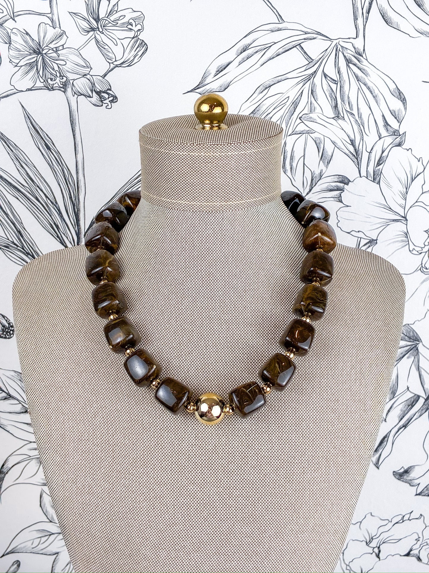 A chunky beaded necklace with dark brown tiger's eye stones and a central gold-toned floral accent, displayed on a beige fabric mannequin bust against a black and white floral wallpaper background.
