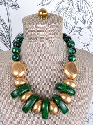 A statement necklace made of alternating polished green beads and chunky gold disc-like beads, displayed on a beige fabric mannequin bust.