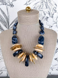 A statement necklace displayed on a beige fabric mannequin bust. The necklace features black marbled stone beads, flat round gold-tone metal discs, and polished light beige wood discs.