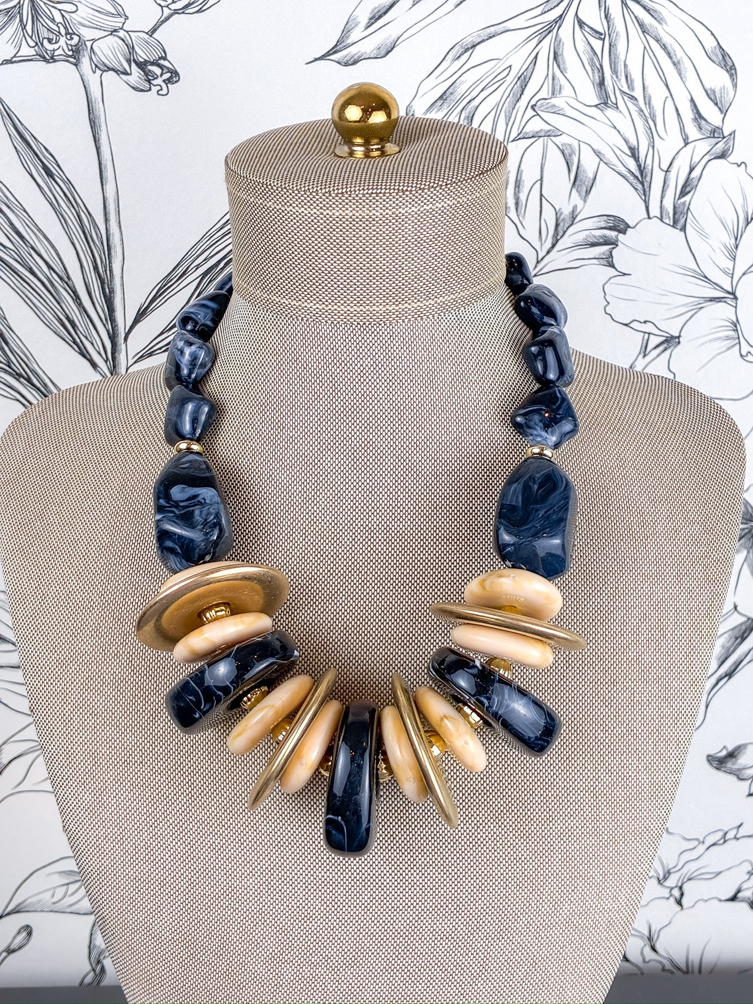 A statement necklace displayed on a beige fabric mannequin bust. The necklace features black marbled stone beads, flat round gold-tone metal discs, and polished light beige wood discs.