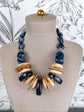 A statement necklace displayed on a beige fabric mannequin bust. The necklace features black marbled stone beads, flat round gold-tone metal discs, and polished light beige wood discs.