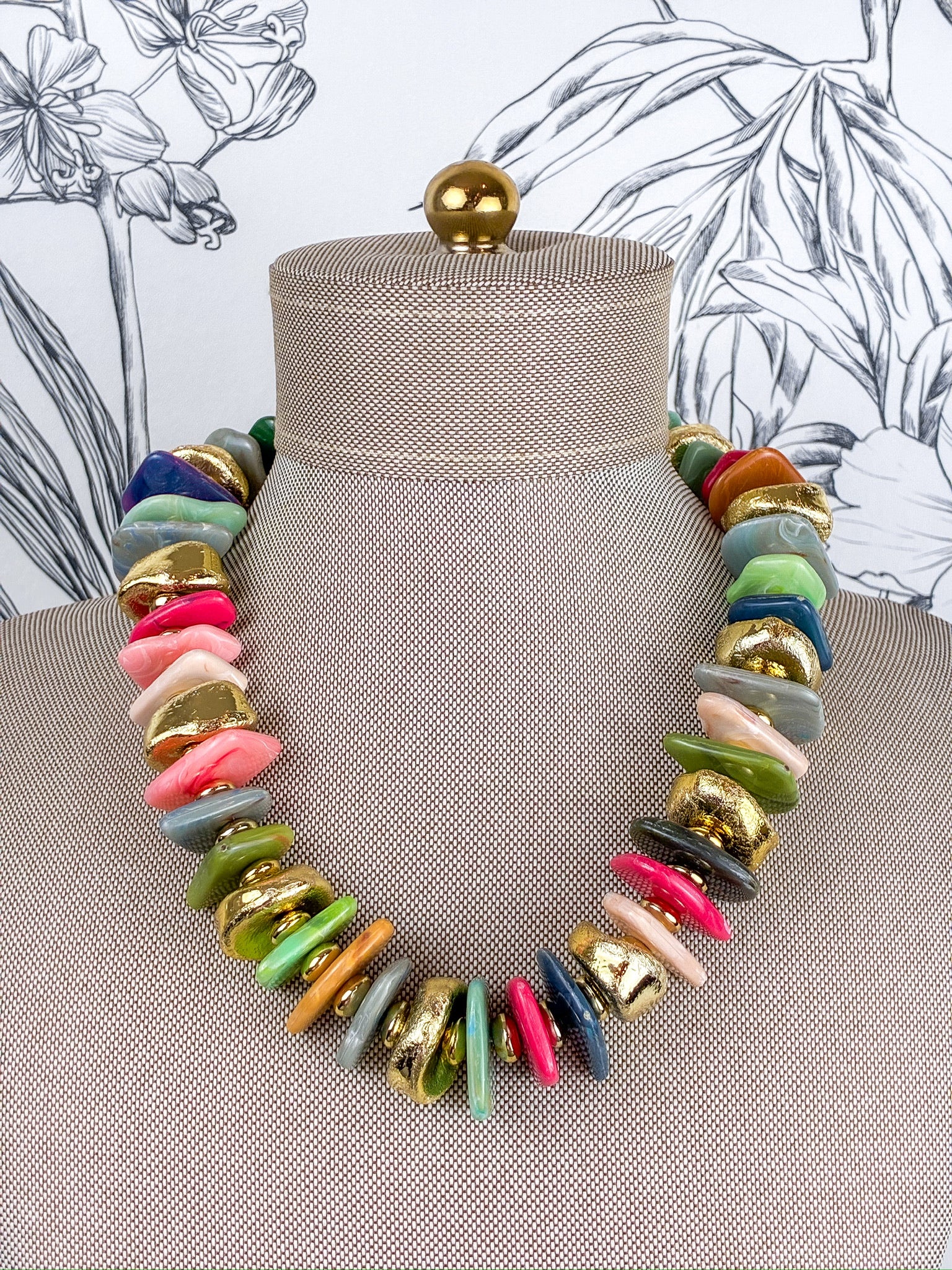 A chunky multi-colored statement necklace made of irregular disc-shaped beads in pink, green, blue, gray, orange, and gold, displayed on a beige fabric mannequin bust.