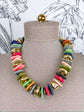 A chunky multi-colored statement necklace made of irregular disc-shaped beads in pink, green, blue, gray, orange, and gold, displayed on a beige fabric mannequin bust.