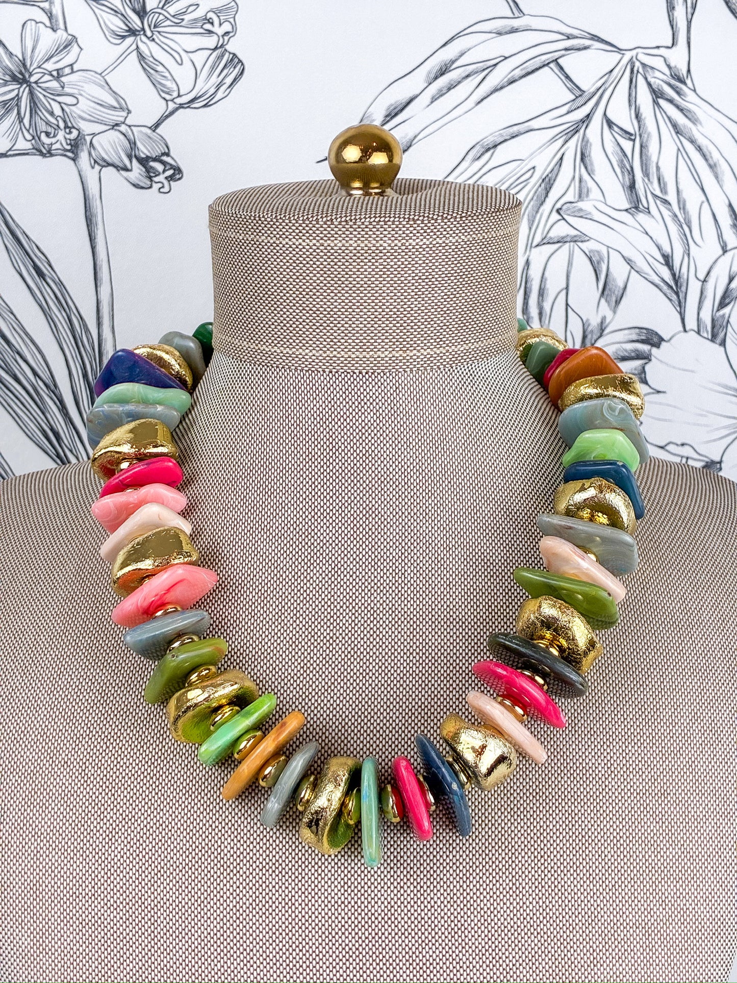 A chunky multi-colored statement necklace made of irregular disc-shaped beads in pink, green, blue, gray, orange, and gold, displayed on a beige fabric mannequin bust.