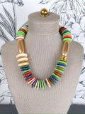 A colorful statement necklace displayed on a beige fabric mannequin bust. The necklace features a mix of flat round beads in green, red, blue, pink, and orange, alongside flat oval beads in beige and metallic gold disc accents.