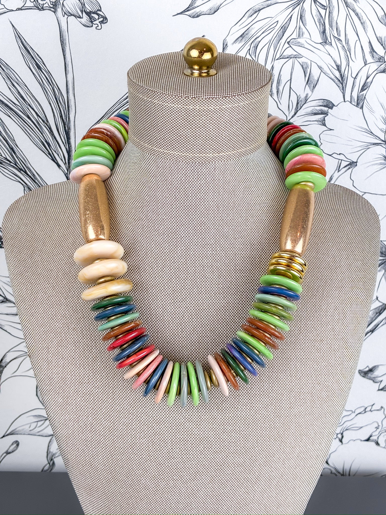 A colorful statement necklace displayed on a beige fabric mannequin bust. The necklace features a mix of flat round beads in green, red, blue, pink, and orange, alongside flat oval beads in beige and metallic gold disc accents.