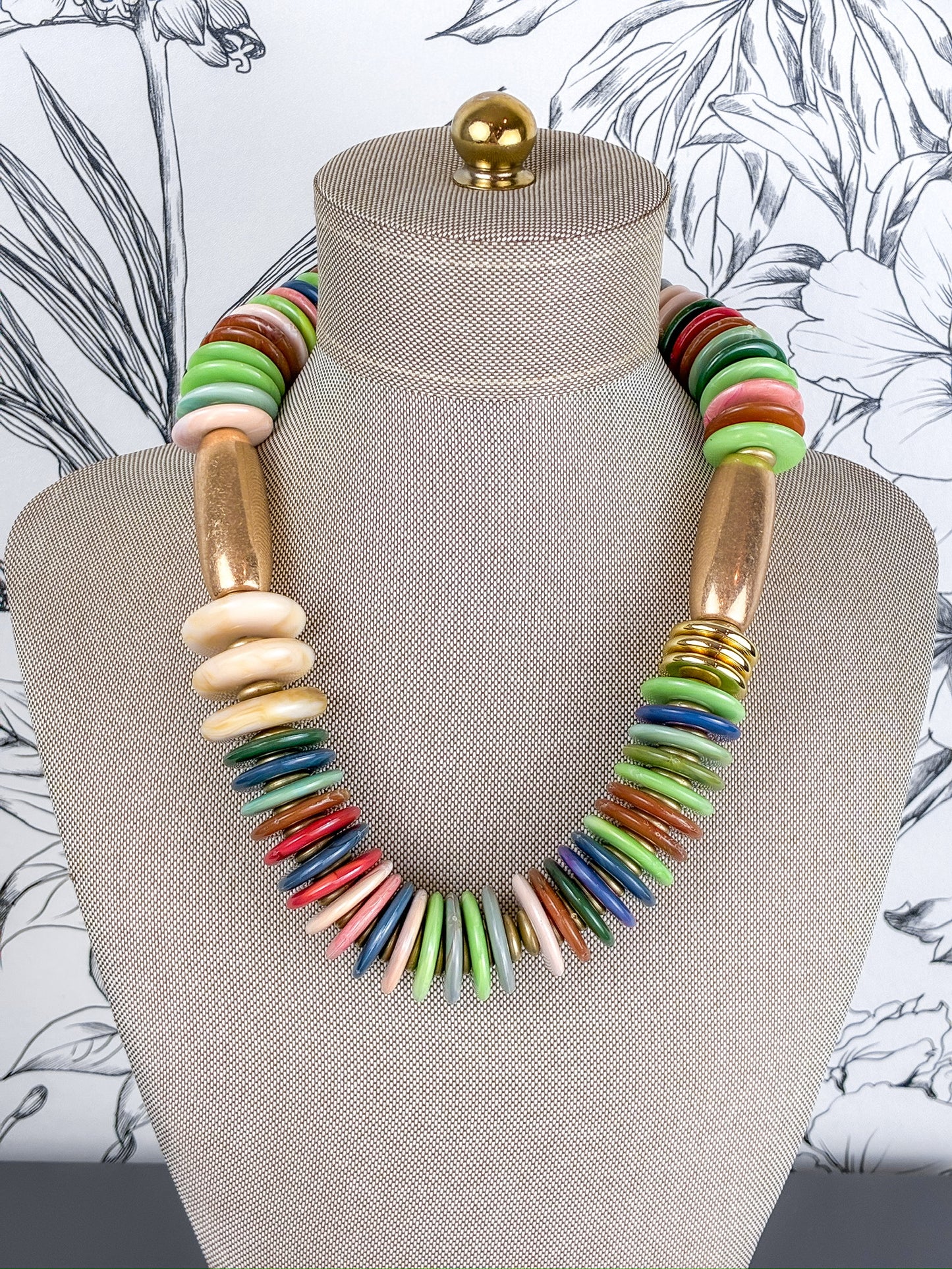 A colorful statement necklace displayed on a beige fabric mannequin bust. The necklace features a mix of flat round beads in green, red, blue, pink, and orange, alongside flat oval beads in beige and metallic gold disc accents.