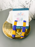 A pair of handmade knotted hair ties in blue and yellow striped fabric, attached to a white display card with a teal header, resting on a gold tray.