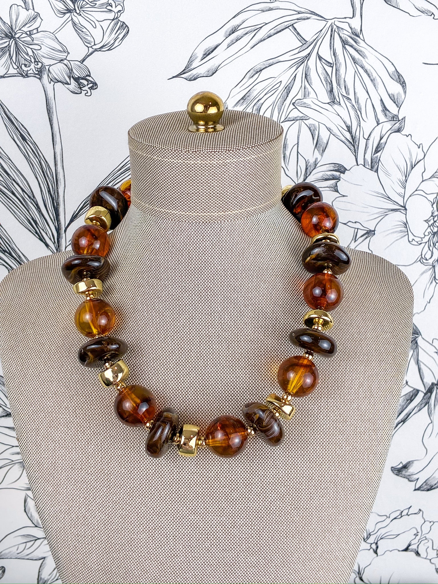 A statement necklace made of polished amber and dark brown glass beads with gold spacer accents, displayed on a beige fabric mannequin bust against a black and white floral wallpaper background.
