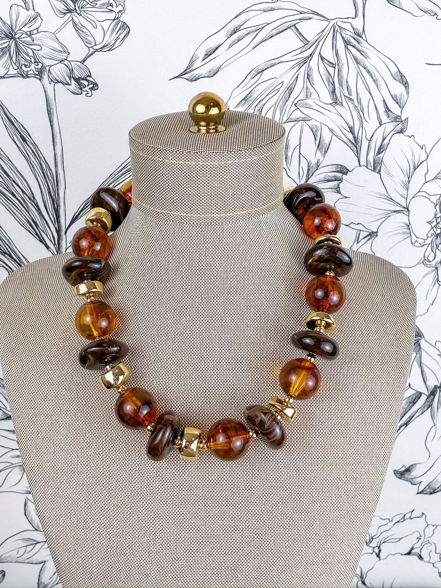 A statement necklace made of polished amber and dark brown glass beads with gold spacer accents, displayed on a beige fabric mannequin bust against a black and white floral wallpaper background.