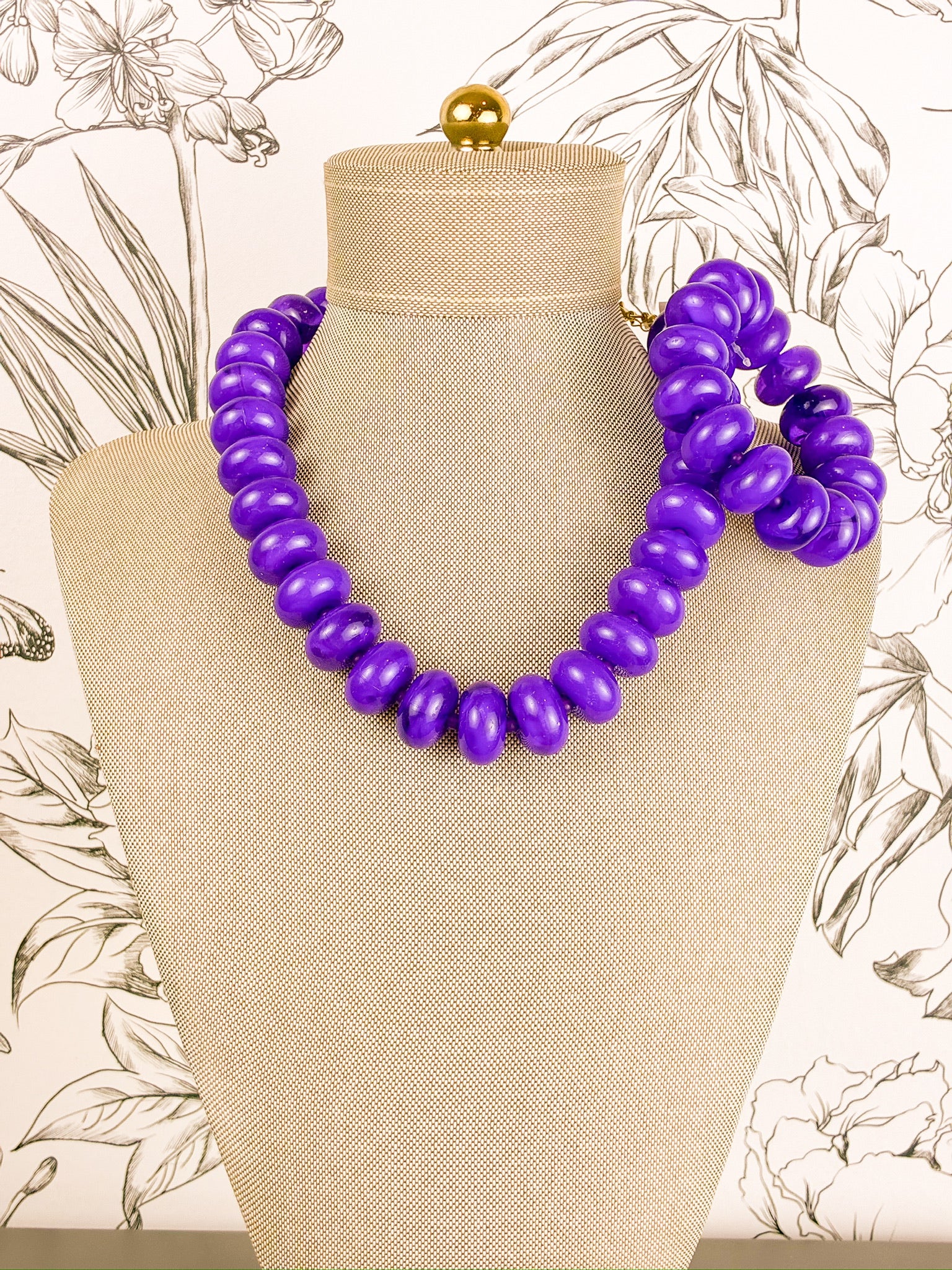 A chunky purple beaded statement necklace set with stretchy bracelet displayed on a beige fabric mannequin bust, set against a light wallpaper with botanical line drawings.