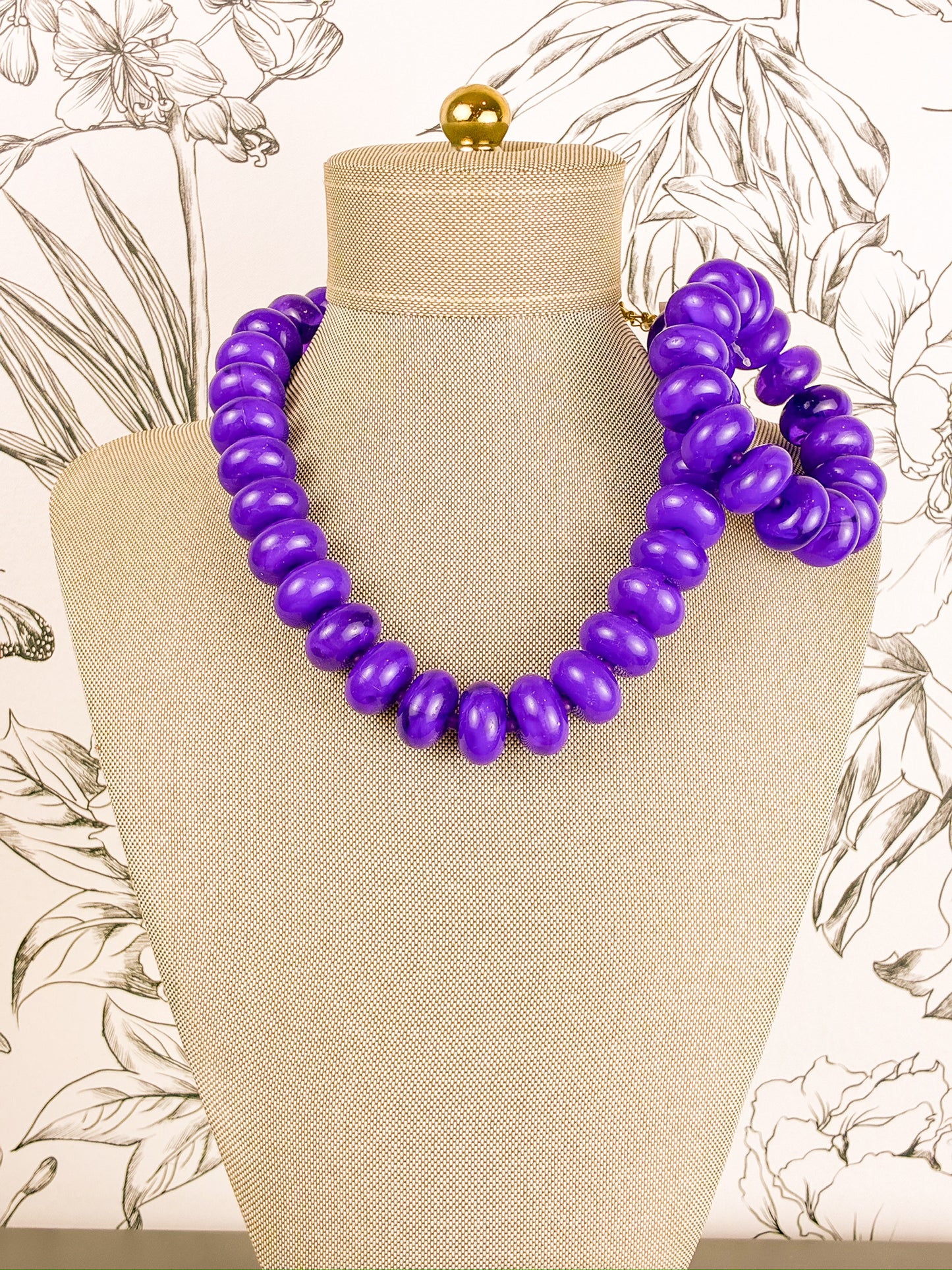 A chunky purple beaded statement necklace set with stretchy bracelet displayed on a beige fabric mannequin bust, set against a light wallpaper with botanical line drawings.