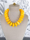 A chunky statement necklace made of glossy, rounded yellow beads, displayed on a beige fabric mannequin bust against a white floral wallpaper background.
