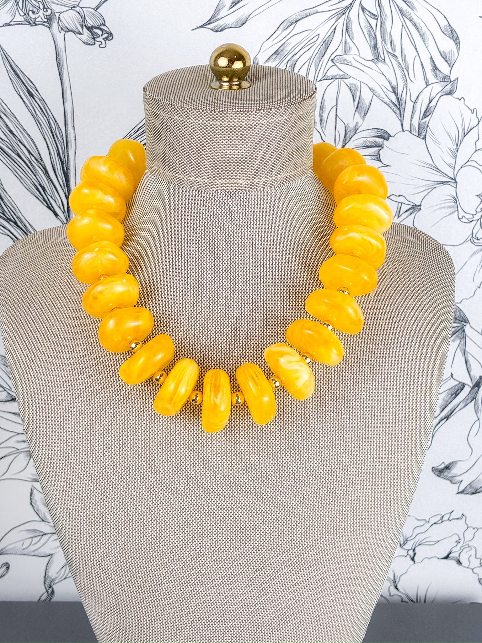 A chunky statement necklace made of glossy, rounded yellow beads, displayed on a beige fabric mannequin bust against a white floral wallpaper background.