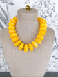 A chunky statement necklace made of glossy, rounded yellow beads, displayed on a beige fabric mannequin bust against a white floral wallpaper background.