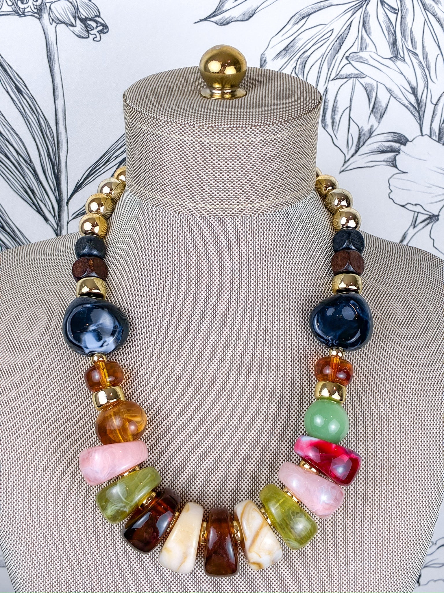 A multicolor statement necklace displayed on a beige fabric jewelry mannequin bust. The necklace features a mix of polished stone beads in dark blue, amber brown, light green, pink, cream, and red, separated by small gold spacer beads.