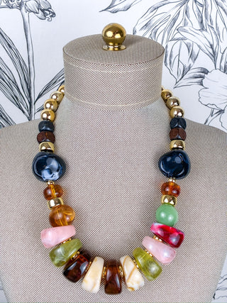 A multicolor statement necklace displayed on a beige fabric jewelry mannequin bust. The necklace features a mix of polished stone beads in dark blue, amber brown, light green, pink, cream, and red, separated by small gold spacer beads.