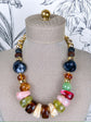 A multicolor statement necklace displayed on a beige fabric jewelry mannequin bust. The necklace features a mix of polished stone beads in dark blue, amber brown, light green, pink, cream, and red, separated by small gold spacer beads.