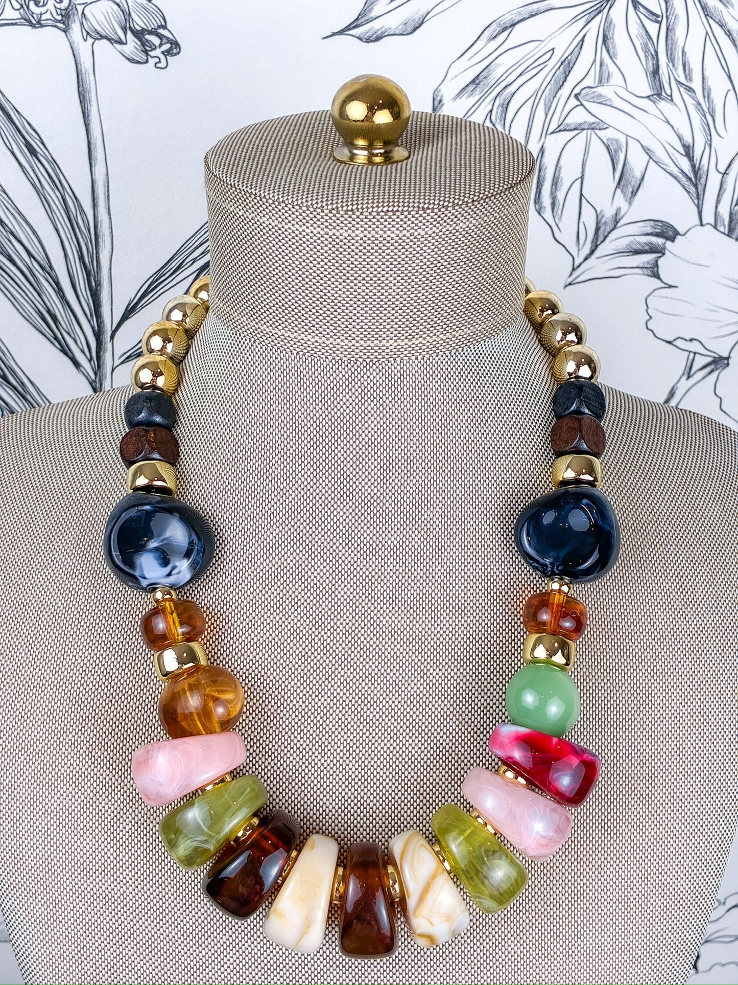 A multicolor statement necklace displayed on a beige fabric jewelry mannequin bust. The necklace features a mix of polished stone beads in dark blue, amber brown, light green, pink, cream, and red, separated by small gold spacer beads.
