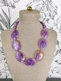 A purple marbled stone bead necklace displayed on a beige fabric-covered jewelry bust against a black-and-white floral background.