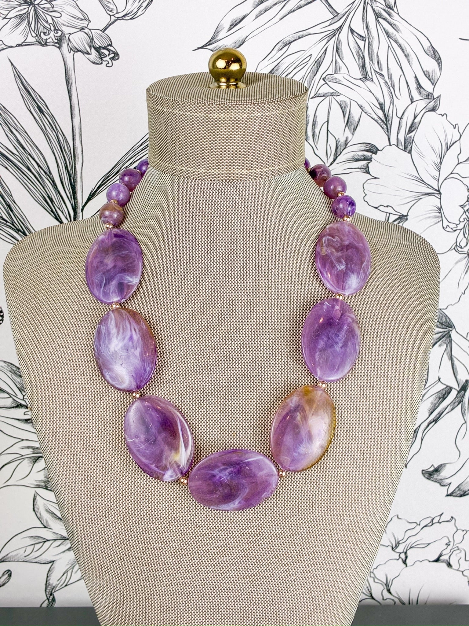 A purple marbled stone bead necklace displayed on a beige fabric-covered jewelry bust against a black-and-white floral background.