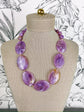 A purple marbled stone bead necklace displayed on a beige fabric-covered jewelry bust against a black-and-white floral background.
